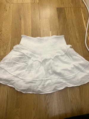 Vit kortkjol med volang från Gina Tricot - Supersöt vit kortkjol från Gina Tricot med bred smock i midjan och volangkant upptill. Kjolen är gjord i ett lätt och luftigt material, perfekt för varma dagar. Passar dig som gillar en feminin och enkel stil.