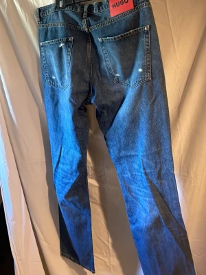 Blå raka jeans från HUGO - Snygga blå jeans från HUGO med raka ben och klassisk femficksdesign. Jeansen har en röd logotyp-patch bak i midjan och subtila slitningar på bakfickorna. Tillverkade i denim för en tidlös look.
