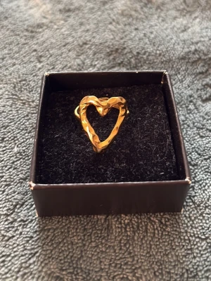 Guldfärgad ring med hjärtform - Säljer en unik guldfärgad ring i metall med öppen hjärtformad design. Ringen har en hamrad yta som ger en cool och trendig look. Perfekt accessoar för dig som gillar statement-smycken och vill sticka ut lite extra.