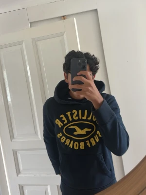 Mörkblå Hollister hoodie med tryck - Snygg mörkblå hoodie från Hollister med stort gult tryck på bröstet där det står 'Hollister Surfboards' och en ikonisk fågel. Hoodien har huva med dragsko och en stor magficka framtill. Perfekt för en avslappnad och cool stil.