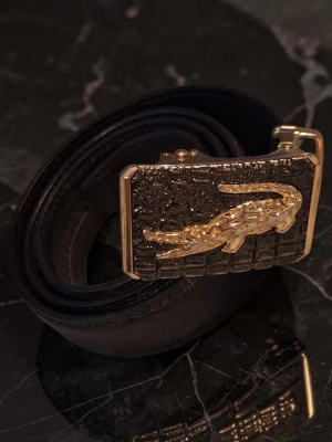 Svart bälte med guldfärgat spänne - Snyggt svart bälte med ett stort, guldfärgat spänne som har en krokodil i relief. Spännet har en texturerad yta som ger en exklusiv känsla. Perfekt accessoar för att lyfta din outfit.