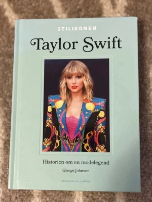 Stilikonen Taylor Swift bok - En inspirerande bok om Taylor Swift, hennes stilresa och ikoniska outfits. Omslaget är ljusblått med ett färgstarkt foto av Taylor i en mönstrad kavaj. Boken är inbunden och skriven av Glenys Johnson. Perfekt för dig som älskar mode, popkultur och vill veta mer om Swifts utveckling.