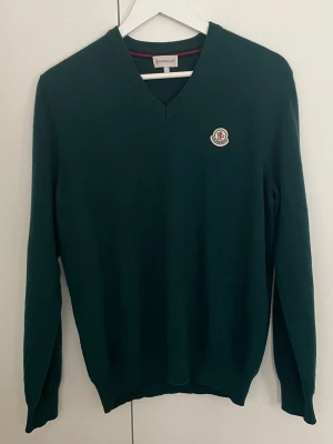 Mörkgrön stickad tröja med V-ringning - Mörkgrön stickad tröja med V-ringning och Moncler-logga på bröstet. Tröjan har långa ärmar och ribbade muddar vid ärmslut och nederkant. Perfekt för dig som gillar stilrena plagg med en exklusiv touch.