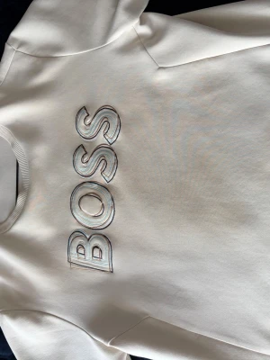 Vit sweatshirt med BOSS-logga - Snygg vit sweatshirt från BOSS med stor broderad logga på bröstet. Tröjan har rund halsringning, långa ärmar och ribbade muddar vid ärmslut och nederkant. Perfekt för en clean och stilren look.