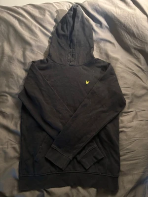 Svart hoodie med gul logga - Säljer en svart hoodie med huva och en liten gul logga på bröstet. Klassisk känguruficka framtill och ribbade muddar vid ärmslut och nederkant. Perfekt för en avslappnad och stilren look.