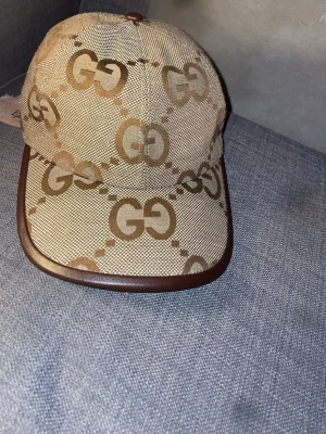 Beige keps med GG-mönster från Gucci - Säljer en beige keps från Gucci med det klassiska GG-mönstret i brunt över hela kepsen. Kepsen har bruna detaljer längs skärmen och är tillverkad i canvasmaterial med insida i ljust tyg. Perfekt för dig som gillar exklusiva accessoarer.