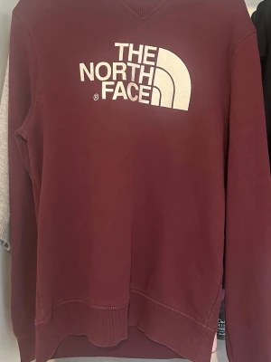 Vinröd sweatshirt från The North Face - Vinröd sweatshirt från The North Face med stor vit logga på bröstet och liten logga på ryggen. Klassisk rund halsringning och ribbade muddar vid ärmslut och nederkant. Perfekt för en avslappnad och sportig stil.
