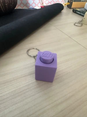 Lila LEGO-nyckelring kub clicker - Säljer en lila nyckelring formad som en stor LEGO-kloss. Tillverkad i plast med tydlig LEGO-logga på toppen. Perfekt att hänga på väskan eller nyckelknippan för en lekfull touch.