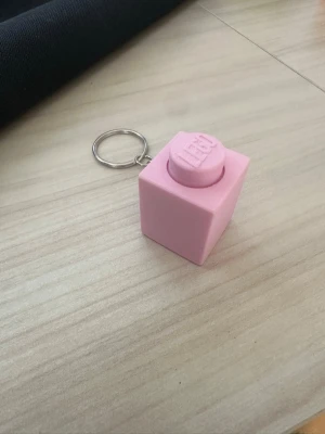 Rosa LEGO-nyckelring kub clicker - Säljer en söt rosa nyckelring formad som en stor LEGO-kloss. Perfekt att hänga på väskan eller nyckelknippan för en lekfull touch. Tillverkad i plast med metallring. LEGO-loggan syns tydligt på toppen av klossen.