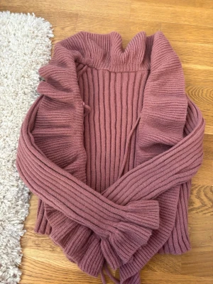 Rosa stickad kofta med volangkant - Supersöt stickad kofta i dammigt rosa med bred volangkant längs hela öppningen. Koftan har ribbstickad struktur och långa ärmar. Perfekt för dig som gillar en romantisk och trendig look. Materialet känns mjukt och mysigt.