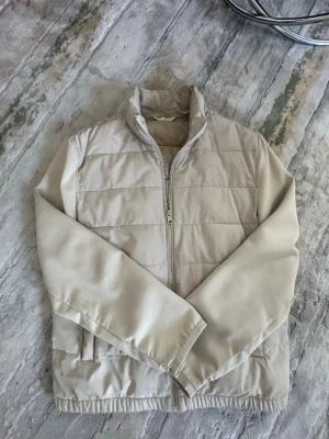 Beige dunväst med dragkedja - Stilren beige dunväst med hög krage och dragkedja framtill. Västen har två sidofickor och quiltad design med horisontella sömmar. Perfekt att slänga över en hoodie eller tröja för extra värme. Materialet är glansigt och ger en modern look. Jag har storlek M i vanligtvis men L passade mig perfekt! 