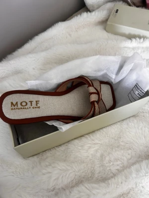 Beige sandaler med knut från MOTF - Snygga sandaler från MOTF i beige tyg med bruna detaljer och en dekorativ knut framtill. Platt sula och öppen tå ger en avslappnad men trendig look. Perfekta för sommaren och lätta att matcha med olika outfits.