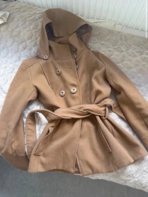 Beige trenchcoat med huva och knytskärp - Stilren beige trenchcoat med huva, dubbelknäppning och knytskärp i midjan. Jackan har fickor på sidorna och en mjuk, något figursydd passform. Perfekt för dig som vill ha en klassisk look med modern twist.