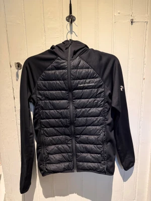 Svart pufferjacka Peak Performance - Snygg svart pufferjacka från Peak Performance med quiltade paneler fram och bak, huva och dragkedja. Jackan har släta ärmar och sidor i stretchigt material för extra komfort. Perfekt för kalla vinterdagar och har en sportig look. Jackan är i nyskick och använd ett fåtal gånger inga slitningar osv. Säljer pga att jag vuxit ur den. Har du frågor är det bara att skriv! Pris går att diskutera 