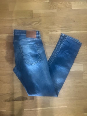 Jack&Jones  - Säljer ett par jack&jones klassiska blå jeans med raka ben och en snygg läderpatch vid linningen. Jeansen har en bakficka och är tillverkade i denim med en tvättad look. Perfekta för dig som gillar en enkel och tidlös stil.