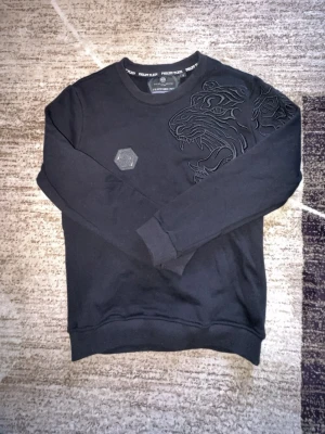 Svart sweatshirt från Philipp Plein - Cool svart sweatshirt från Philipp Plein med ett stort broderat tigerhuvud på bröstet och en sexkantig logodetalj. Tröjan har rund halsringning och ribbade muddar vid ärmslut och nederkant. Perfekt för dig som gillar streetwear och detaljer.