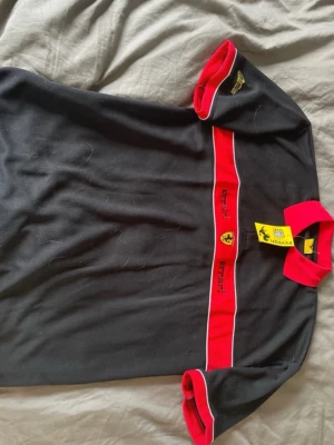 Svart skjorta med röda Ferrari-detaljer - Snygg vintage svart polo med röda detaljer och vit kant. Framtill finns en bred röd rand med Ferrari-logga och broderad text. Kragen och ärmsluten är också röda, vilket ger en sportig look. Perfekt för dig som gillar bilar och vill sticka ut. Storlek XL/L, gamla prislappen kvar