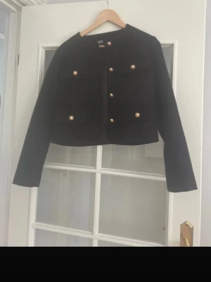 Svart kort kavaj med guldknappar - Stilren svart kavaj i kort modell med raka axlar och fyra framfickor. Kavajen har dekorativa guldknappar framtill och på fickorna. Passar perfekt till både jeans och kjol för en trendig look. Materialet ser ut att vara en mjuk och stadig textil.