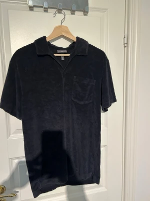 Svart frottéskjorta från H&M - Säljer en svart kortärmad skjorta från H&M i mjuk frotté. Skjortan har klassisk krage, bröstficka och relaxed fit. Perfekt för en chill och avslappnad stil. Regular fit-modell.