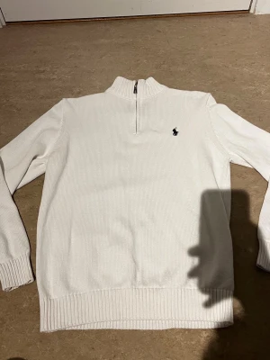 Vit stickad tröja med halv dragkedja - Vit stickad tröja från Polo Ralph Lauren med halv dragkedja och ribbade muddar. Klassisk broderad logga på bröstet i svart. Tröjan har hög krage och är tillverkad i ett mjukt, tjockt material som passar perfekt för kyligare dagar.