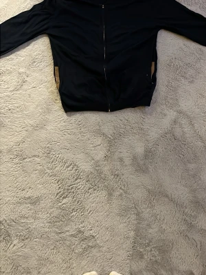 Svart hoodie med mockadetaljer - Svart hoodie med dragkedja och bruna mockadetaljer på armbågar och vid fickor. Klassisk passform med långa ärmar och två sidofickor. Perfekt för en chill och stilren look.