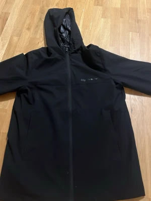 Svart regnjacka från Jack & Jones - Svart regnjacka från Jack & Jones med huva och hel dragkedja framtill. Jackan har två sidofickor och är tillverkad i ett vattenavvisande material med slät finish. Perfekt för regniga dagar och enkel att matcha med olika outfits.