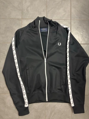 Fred Perry tracktop - nyskick - Snygg svart tracktop från Fred Perry med vita detaljer och dragkedja. Tracktopen är i toppskick och har endast använts vid 3 tillfällen. Storlek S Tröjan köptes hos Thernlunds för ca 1300kr, jag säljer en i princip helt ny här för 700kr. Skicka PM för ytterligare bilder eller frågor!