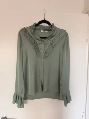 Grön volangblus från NA-KD - Stilren ljusgrön blus från NA-KD med volangdetaljer längs krage och bröst. Blusen har knäppning framtill och långa ärmar med volang vid ärmslut. Tillverkad i ett glansigt, mjukt material som ger en elegant känsla.