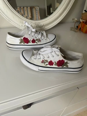 Vita sneakers från pimkie  med röda rosbroderier - Säljer ett par pimkie vita låga sneakers med snygga broderade röda rosor och gröna blad på sidorna. Skorna har vita snören, svart rand runt sulan och klassisk gummitå. Perfekta för dig som vill ha en unik och trendig look.storlek är precis som en 38.kan möta er ,så slipper ni frakt 