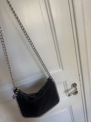 Svart axelväska med kedjedetaljer - Cool svart axelväska i fejkläder med grova silverfärgade kedjor runt kanterna och som axelrem. Väskan har en mjuk, lite slouchy form och dragkedja på baksidan. Perfekt för dig som gillar edgy detaljer och vill sticka ut. Litet slitage vid magneten, annars bra skick. 