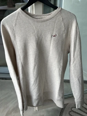 Beige tröja från Hollister - Snygg beige tröja från Hollister med rund halsringning och diskret vinröd logga broderad på bröstet. Tröjan har långa ärmar och ribbade muddar vid ärmslut och nederkant. Perfekt för en clean och avslappnad stil.