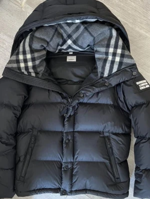 Burrberry väst/jacka - Snygg svart pufferjacka med stor huva och rutigt foder i grått och vitt. Jackan har dragkedja och knappar framtill, två snedställda fickor med dragkedja och en patch på ärmen. Puffig modell som håller värmen och ger en cool look.