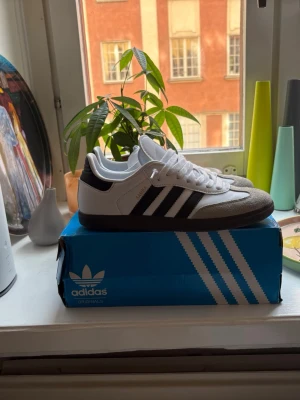 Adidas Samba  - Adidas Samba OG sneakers i vitt och svart med klassiska tre ränder på sidan, grå mockadetaljer vid tån och svart häl. Ovandelen är i läder och mocka, yttersulan är i gummi. Snygg retrostil som funkar till det mesta. Skorna är 40,5 men sitter som 41