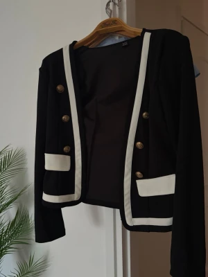 Svart kavaj med vita detaljer - Stilren svart kavaj med vita kontrastkanter och dekorativa guldknappar framtill. Kavajen har två fickor med vita lock och raka axlar. Passar perfekt till en trendig och elegant look.
