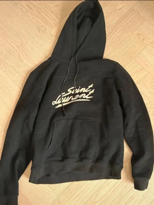 Svart Saint Laurent hoodie med tryck - Säljer en svart hoodie från Saint Laurent med vit logga i graffiti-stil på bröstet. Hoodien har huva med dragsko och en stor magficka. Tillverkad i mjukt bomullsmaterial och har ribbade muddar vid ärmar och nederkant.