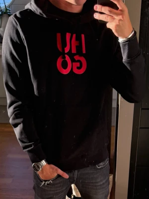 Svart hoodie från HUGO med röd logga - Snygg svart hoodie från HUGO med stor röd logga på bröstet. Klassisk huva med dragsnören och en stor magficka framtill. Tillverkad i mjukt material som känns skönt mot huden. Perfekt för en avslappnad och trendig look.