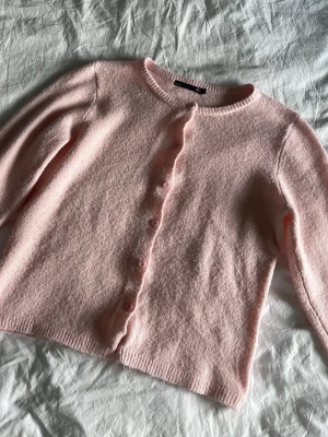 Ljusrosa kofta från 157 - Mysig ljusrosa kofta från 157 med rund halsringning och knappar framtill. Koftan har lång ärm och är stickad i ett mjukt material som känns skönt mot huden. Perfekt att slänga över en topp för en söt och enkel look.