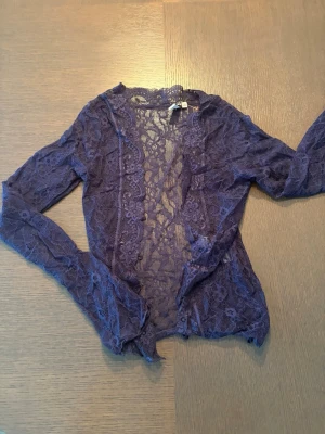 Lila spetsbolero med lång ärm - Säljer en mörklila bolerotopp i genomskinlig spets med blommönster och dekorativa kanter. Toppen har lång ärm och är perfekt för att lägga till en extra detalj till din outfit.
