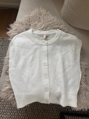 Vit ärmlös kofta med knappar - Stilren vit ärmlös kofta med rund halsringning och knäppning framtill. Koftan har ribbade kanter vid hals och nederkant, vilket ger en snygg finish. Perfekt att bära över en t-shirt eller skjorta för en trendig lager-på-lager-look.🤍
