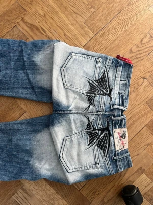 Blå jeans med broderade vingar - Säljer ett par blå jeans med coola svarta broderade vingar på bakfickorna. Jeansen har en tvättad look med ljusare partier och raka ben. Materialet är klassisk denim och de har två bakfickor. Midjemåttet är 36 cm rakt över och Innerbenslängden 82 cm.