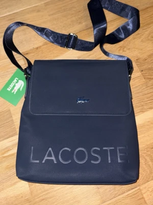 Mörkblå axelväska från Lacoste - Snygg mörkblå axelväska från Lacoste med justerbar axelrem och präglad logga framtill. Väskan har ett lock med metallfärgad krokodil och är tillverkad i slitstarkt tygmaterial. Perfekt för att bära det viktigaste med stil.