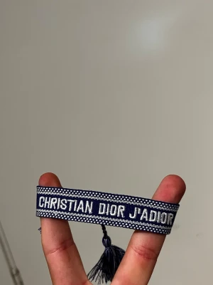 Blått Christian Dior textarmband - Snyggt blått tygarmband från Christian Dior med texten 'CHRISTIAN DIOR J'ADIOR' i vitt. Armbandet har dekorativa tofsar i ändarna och ett vävt mönster längs kanterna. Perfekt accessoar för att lyfta din outfit.