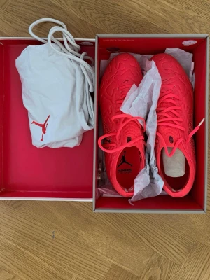 Röda fotbollsskor från Jordan - Snygga röda fotbollsskor från Jordan med klassisk Jumpman-logga. Skorna har en modern, strukturerad ovandel och levereras med en vit dustbag och extra guldfärgade dragkedjor. Perfekta för dig som vill sticka ut på planen.