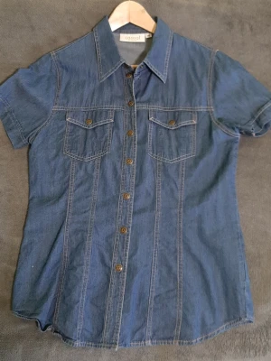 Blå jeansskjorta med kort ärm - Snygg blå jeansskjorta i bomull med korta ärmar och klassisk krage. Skjortan har knappar framtill och två bröstfickor med lock. Perfekt för en avslappnad och trendig look.