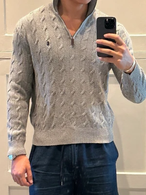 Beige stickad tröja Polo Ralph Lauren - Säljer en beige stickad tröja från Polo Ralph Lauren med klassiskt kabelstickat mönster och half zip-dragkedja. Tröjan har hög krage, långärmad passform och den ikoniska Polo-loggan broderad på bröstet. Perfekt för dig som gillar stilren och tidlös design.