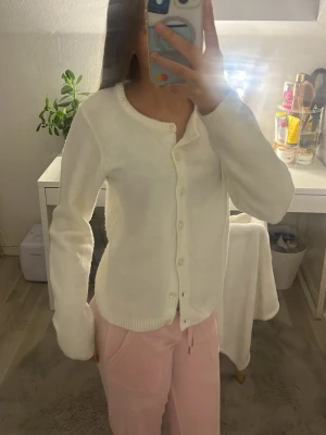 Vit kofta från 157, rund hals - Mysig vit kofta från 157 med rund halsringning och knäppning framtill. Koftan har långa ärmar och ribbade muddar vid ärmslut och nederkant. Perfekt att slänga över en t-shirt eller linne för en clean och enkel look.
