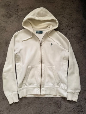 Vintage Polo Ralph Lauren - Säljer en stilren vit hoodie från Polo Ralph Lauren med dragkedja framtill och klassisk broderad logga på bröstet. Hoodien har kängurufickor, ribbade muddar och en mjuk insida. Perfekt för en avslappnad look.