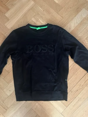 Svart sweatshirt från Hugo Boss - Snygg svart sweatshirt från Hugo Boss med stor BOSS-logga broderad på bröstet. Tröjan har rund halsringning, långa ärmar och ribbade muddar vid ärmslut och nederkant. Tillverkad i mjukt material för en bekväm känsla. Perfekt för en stilren och avslappnad look.