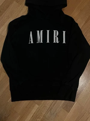 Svart hoodie från Amiri - Säljer en svart hoodie från Amiri med stor vit logga framtill. Klassisk passform med huva och känguruficka. Tillverkad i mjukt material som känns skönt mot huden. Perfekt för dig som gillar streetwear och vill ha en stilren look.