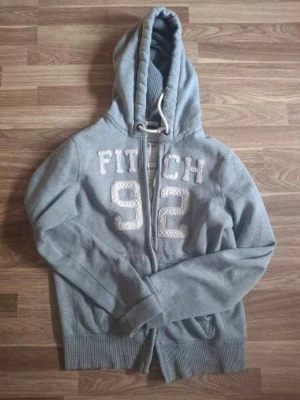 Abercrombie & Fitch Zip Hoodie - Snygg grå hoodie med dragkedja och huva. På bröstet står det 'FITCH 92' i stora vita bokstäver. Hoodien har ribbade muddar och fickor framtill. Perfekt för en avslappnad och sportig stil.